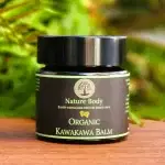 Container of Nature Body Kawakawa Balm 30g on a wood table with some greenery behind it.