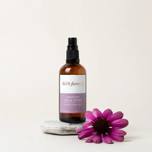 HerbFarm Renew Refine Facial Toner 100ml spray bottle sitting on a rock next to a purple flower