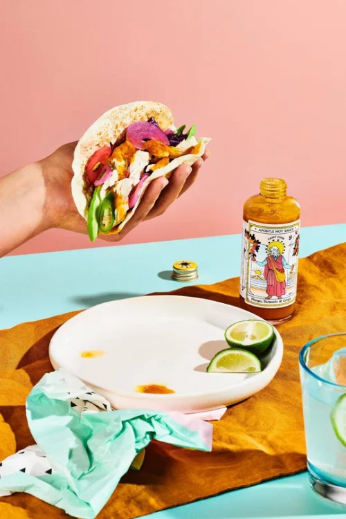 Apostle Saint John Mango Turmeric and Ginger 150ml on a table with a hange holding a taco over a white plate with drippings from the taco on it. There is also a cut up lime on the plate as well. This is next to a glass of margarita. The table is light blue and the background is pink