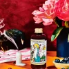 Apostle St Andrew Szechuan Blueberry and Habanero 150ml on a table with a smoking candle, pink flowers in a blue vase, burnt matches, bowl of blueberries, dried up flower, pink and white striped cloth, with a red velvet background