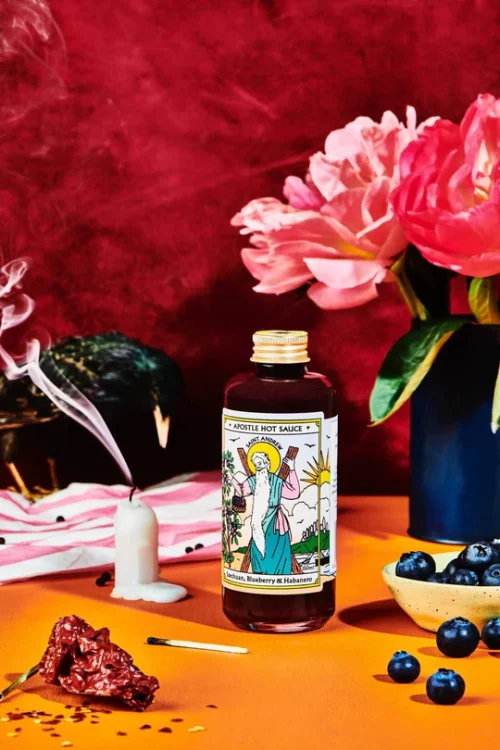 Apostle St Andrew Szechuan Blueberry and Habanero 150ml on a table with a smoking candle, pink flowers in a blue vase, burnt matches, bowl of blueberries, dried up flower, pink and white striped cloth, with a red velvet background