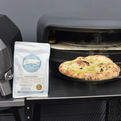 OrganicDough Pizza Dough Ball cooked in front of a pizza oven, next to its packaging