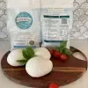 3 pizza dough balls on a round wooden cutting board with tomatoes and basil. There are two of the dough's packaging behind.