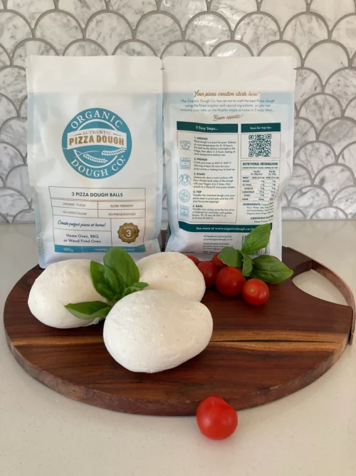 3 pizza dough balls on a round wooden cutting board with tomatoes and basil. There are two of the dough's packaging behind.