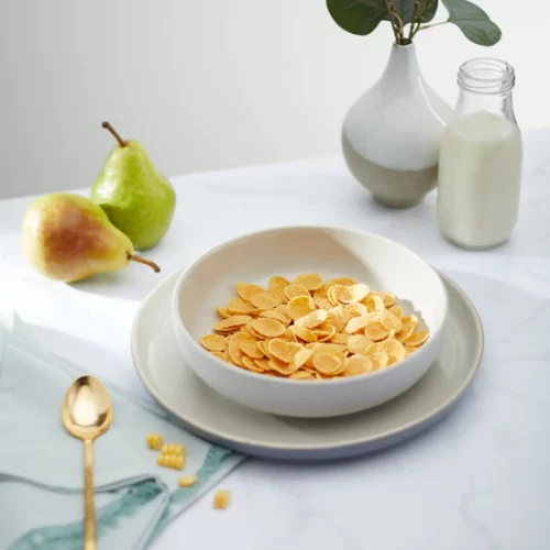 a bowl of NaturesPath Corn Flakes next to pears, a jar of milk, and a green plant in a vase