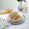 a bowl of NaturesPath Heritage Flakes on a table next to barley, a jar of milk, and a green plant in a vase.